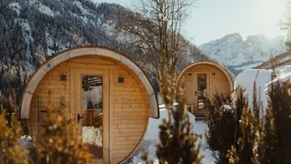 Zwei hölzerne fassförmige Saunen in einer verschneiten Berglandschaft.